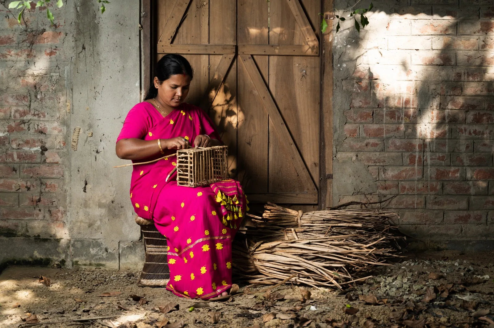 An Asama artisan in a pink mekhela weaving a basket frame at the workshop door in Alimur