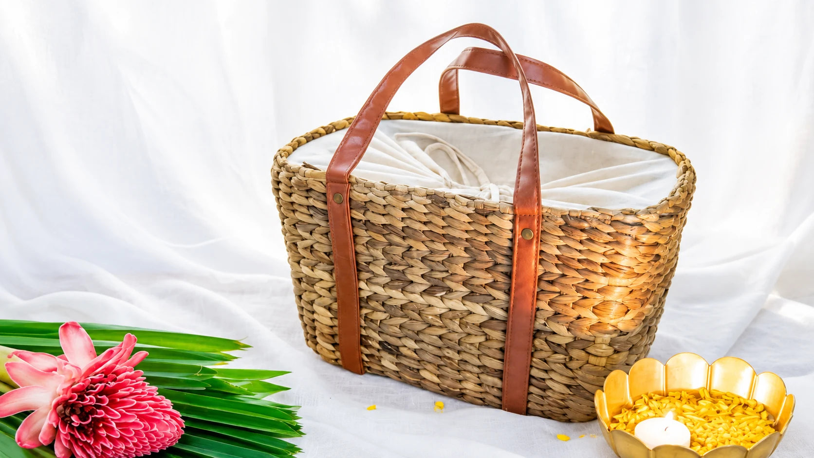 A water hyacinth hamper basket with leather straps and a cotton lining, set beside brass diyas